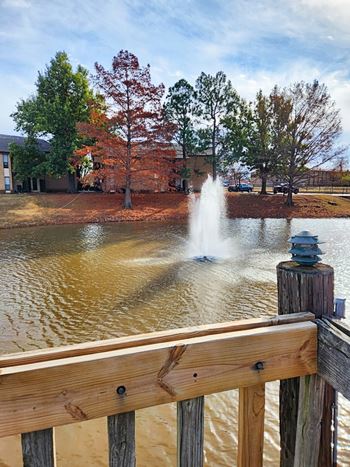 A fountain in the middle of a pond with a wooden fence in the foreground.